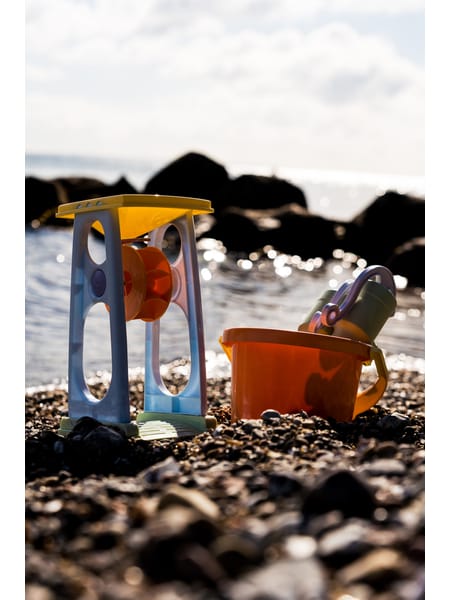 Gul og blå sandmølle med rød spand på strand med sten.